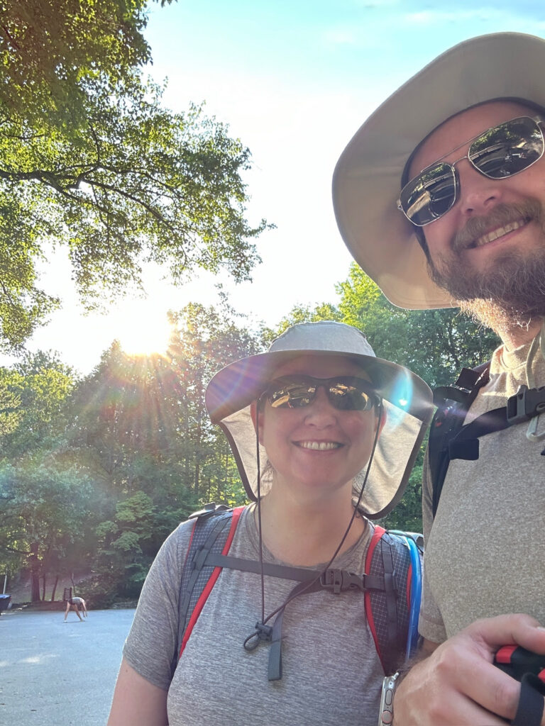 Man and woman pose for a selfie on a summer time hike. Both are wearing a hat and sun glasses because the sun is bright. 